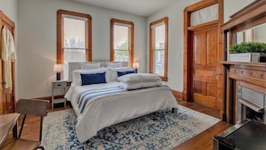 A bedroom with a bed, nightstands, a chair, a wooden door, and a fireplace. - The Baker (North Little Rock)