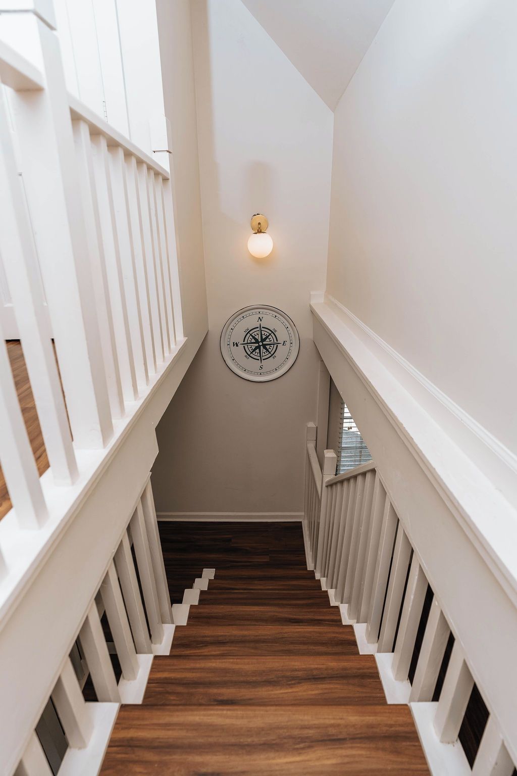 Un escalier avec des rampes blanches et un plancher en bois, menant à une pièce dotée d’un luminaire fixé au mur et décorée d’une boussole.
