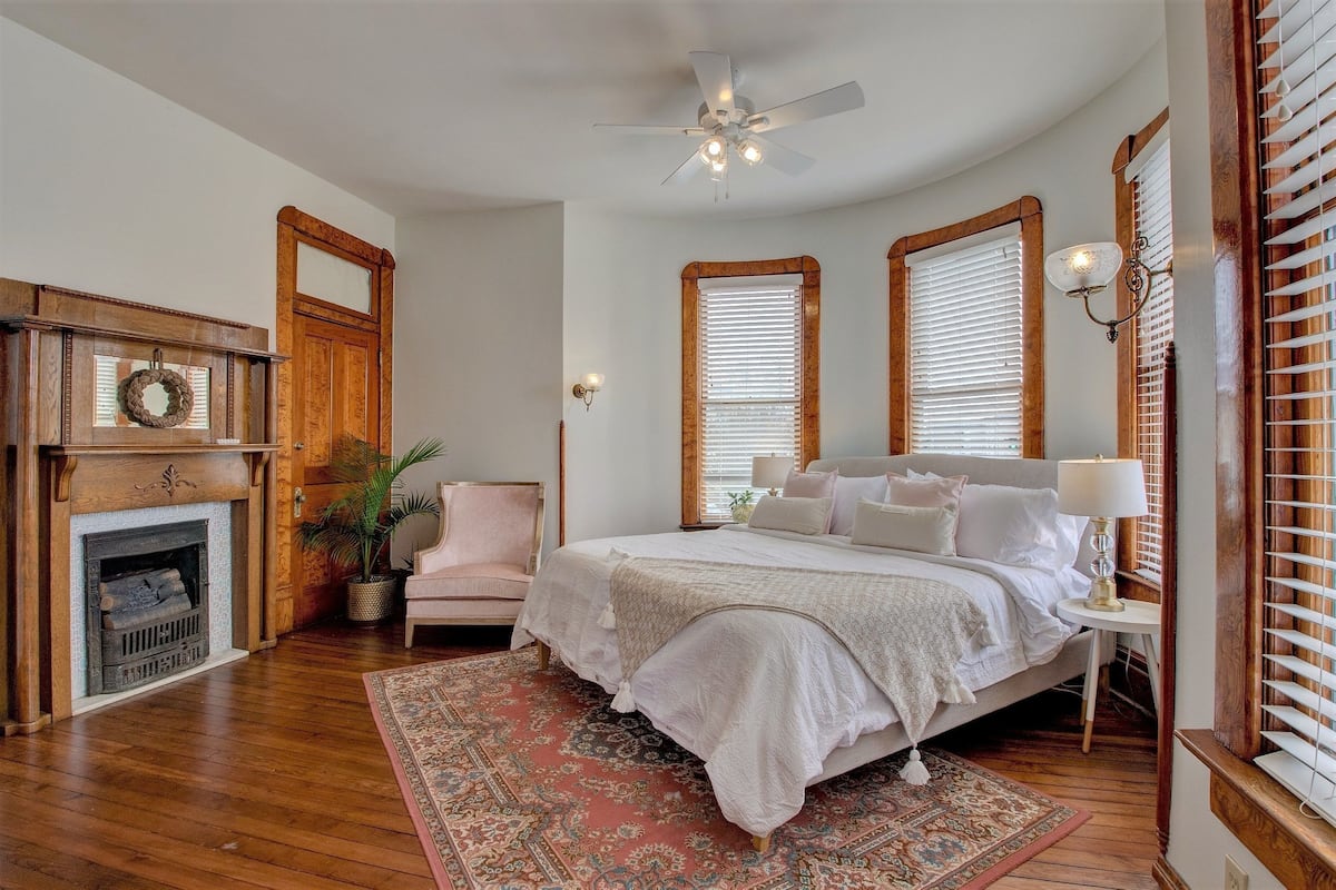 A bedroom with a large bed, a fireplace, a ceiling fan, and wooden window frames.