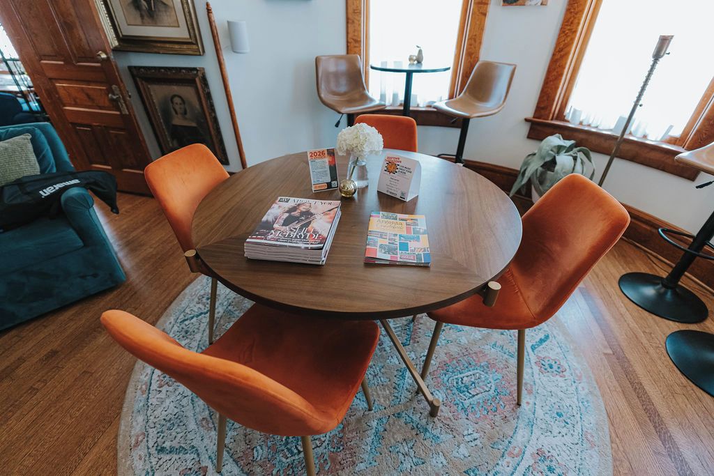 Una mesa redonda de madera con sillas anaranjadas, un sofá azul y un tapete estampado en una habitación luminosa con ventanas grandes.