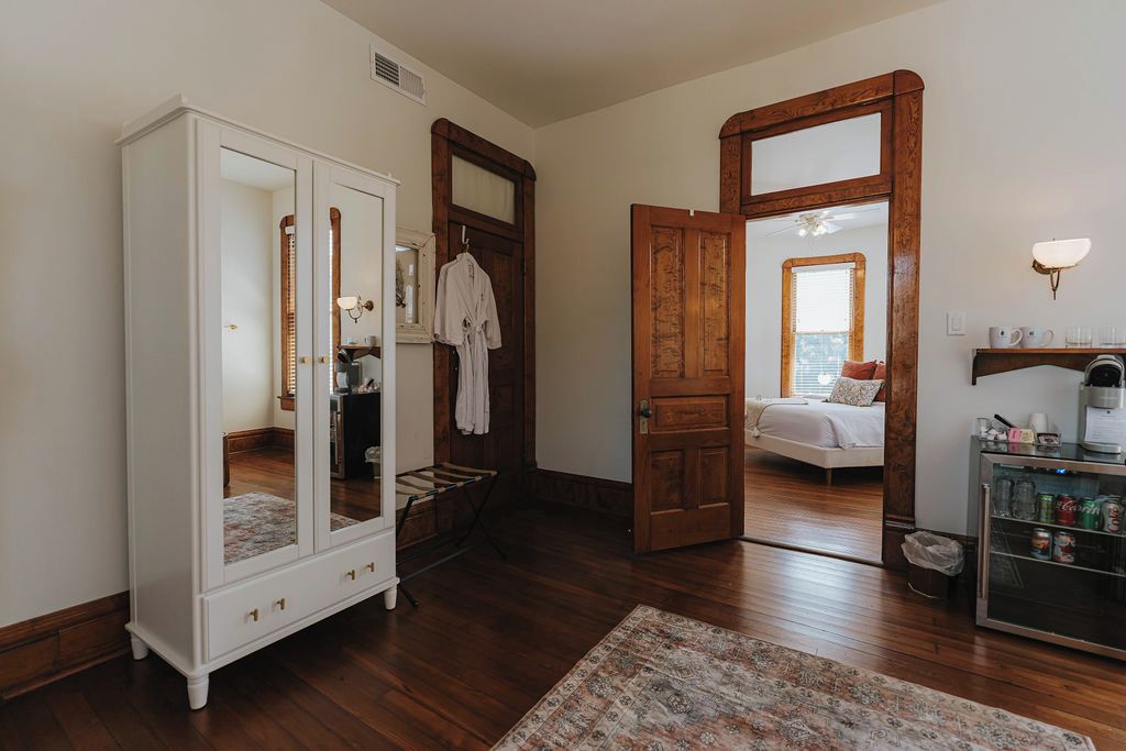 A room with a wooden wardrobe, a bed, a mirror, and a small cabinet with bottles.