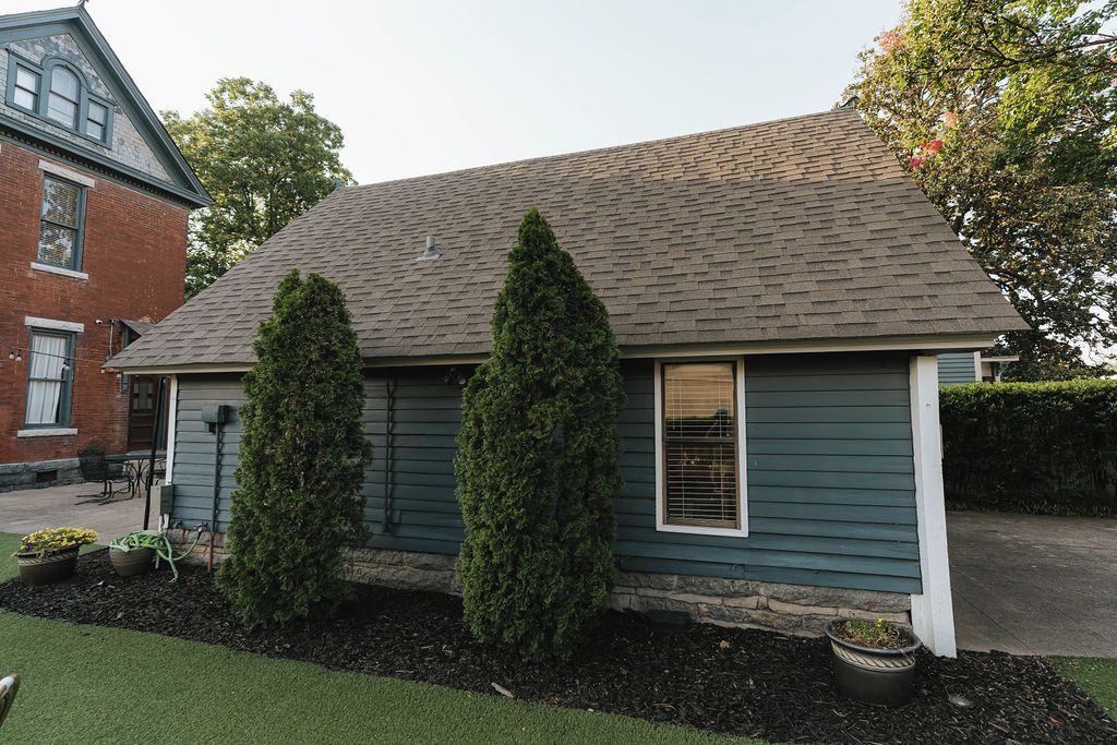 A two-story house with a dark green exterior, a sloped roof, and two tall evergreen trees in front.