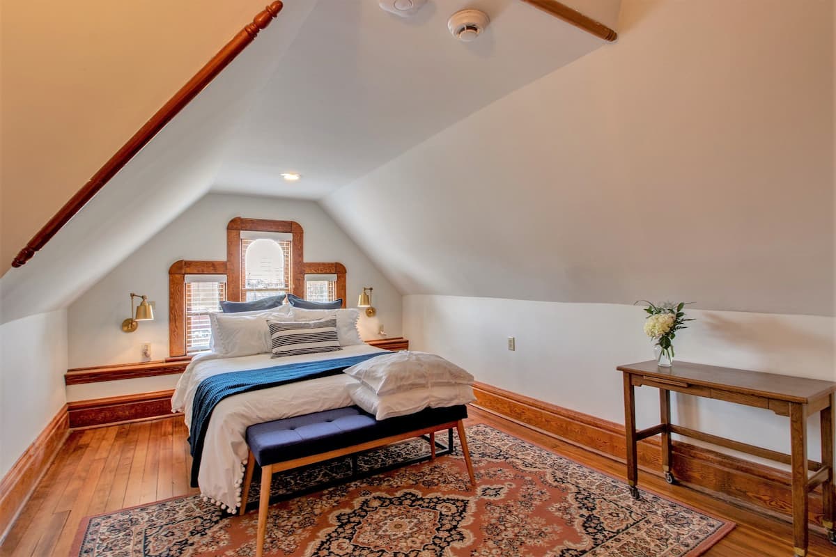 A bedroom with a sloped ceiling, a wooden bench, a bedside table, a patterned rug, and a small table with a vase.