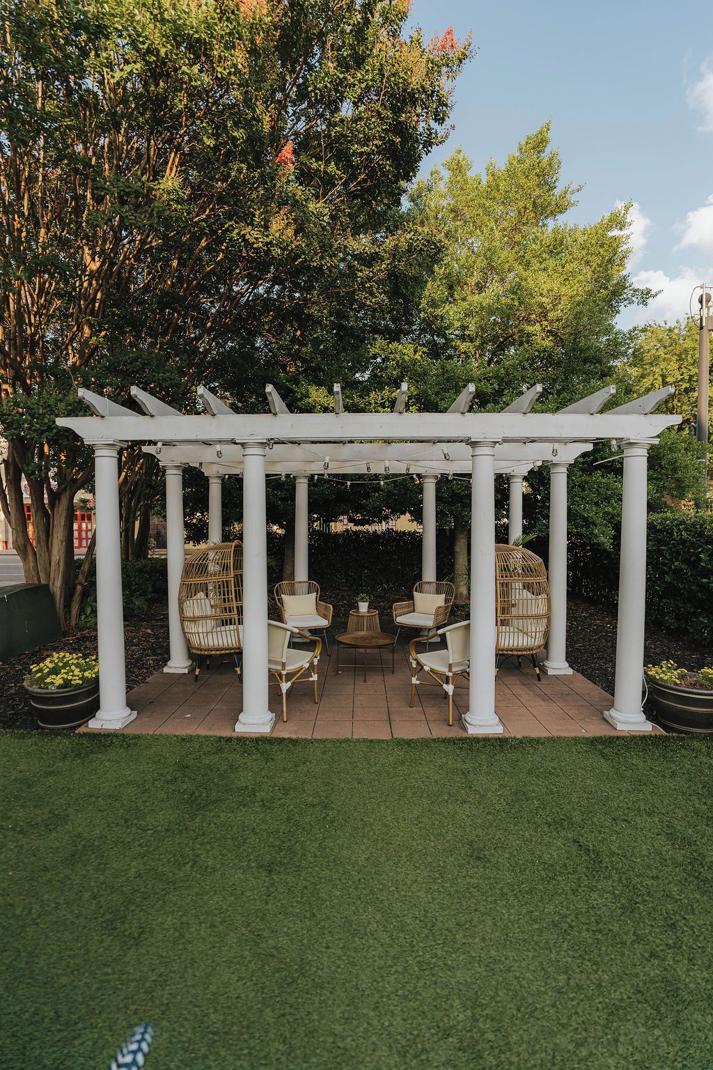 Una estructura de pérgola blanca con cuatro columnas, rodeada de sillas de mimbre y una mesa redonda pequeña, ubicada en un patio empedrado con césped al frente.