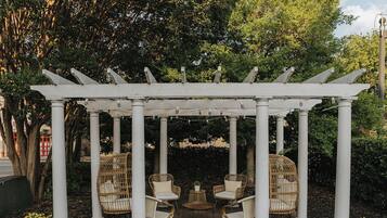Una estructura de pérgola blanca con cuatro columnas, rodeada de sillas de mimbre y una mesa redonda pequeña, ubicada en un patio empedrado con césped al frente.
