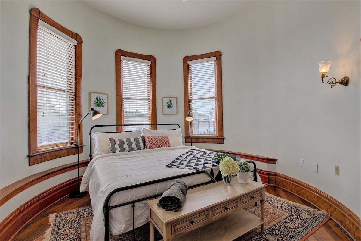A bedroom with a bed, wooden trim, a nightstand, a lamp, and a window with blinds.