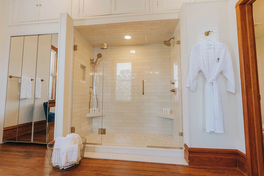 A bathroom with a glass-enclosed shower, a wooden floor, a white robe hanging on the wall, and a mirrored wardrobe.