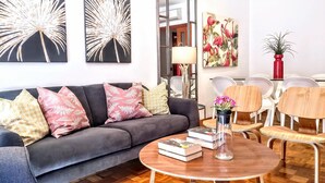 Un salon moderne avec un canapé gris, une table basse ronde en bois agrémentée de livres et d’un vase, et un coin repas avec des chaises blanches et un vase rouge.