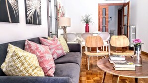 Un salon avec un canapé gris, des coussins à motifs, une table basse ronde sur laquelle sont posés des livres, et un vase rouge contenant des fleurs.