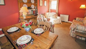 A cozy living room with a wooden dining table set for a meal, a floral sofa, and a bookshelf filled with books and decorative plates.