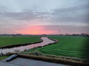 A rural landscape with a winding canal, green fields, and a wind turbine in the distance. - Apartment with a sweeping view of beautiful sunsets (Dornumersiel)