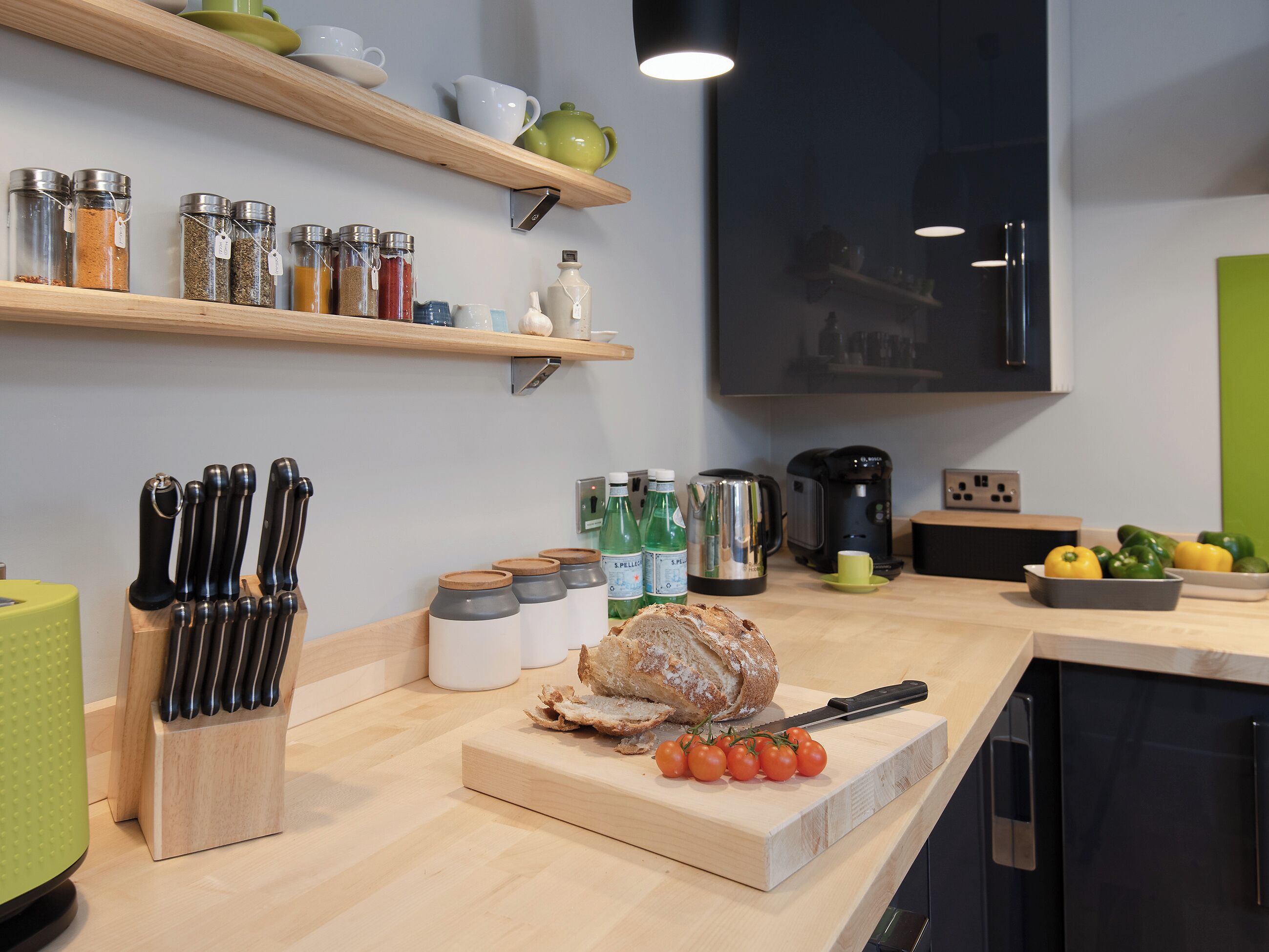 Une cuisine avec des étagères en bois, une planche à découper avec des tomates, une miche de pain et divers ustensiles de cuisine.