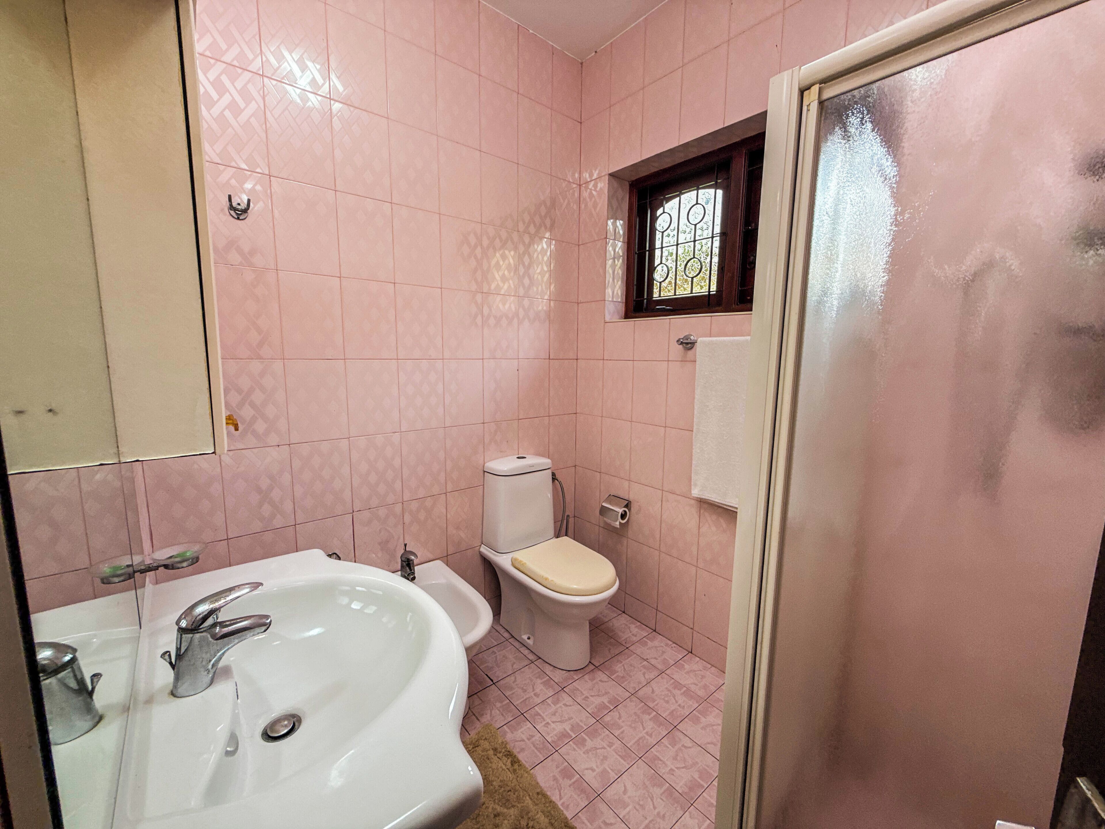 Een badkamer met roze betegelde wanden, een wit wastafelmeubel met een moderne kraan, een toilet en een klein raam met decoratief rooster.