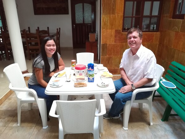 Un comedor con una mesa preparada para dos, sillas de plástico blancas y un banco verde.
