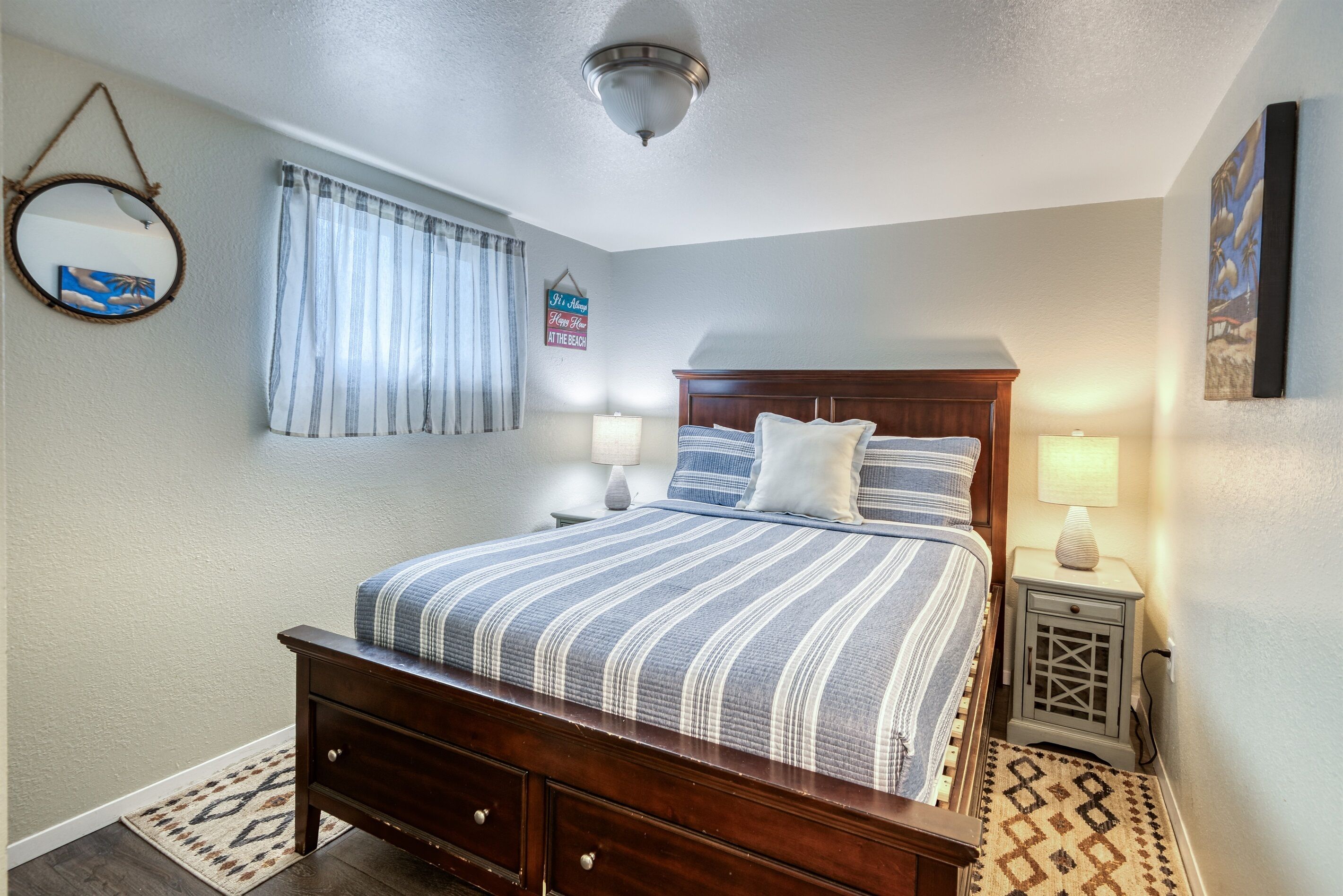 Un dormitorio ordenado con una cama de madera, dos mesitas de noche con lámparas, un espejo en la pared y un letrero con motivos playeros.