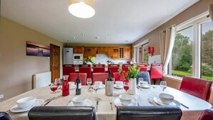 A dining area with a table set for four, surrounded by red chairs, a kitchen in the background, and a view of greenery through large windows.