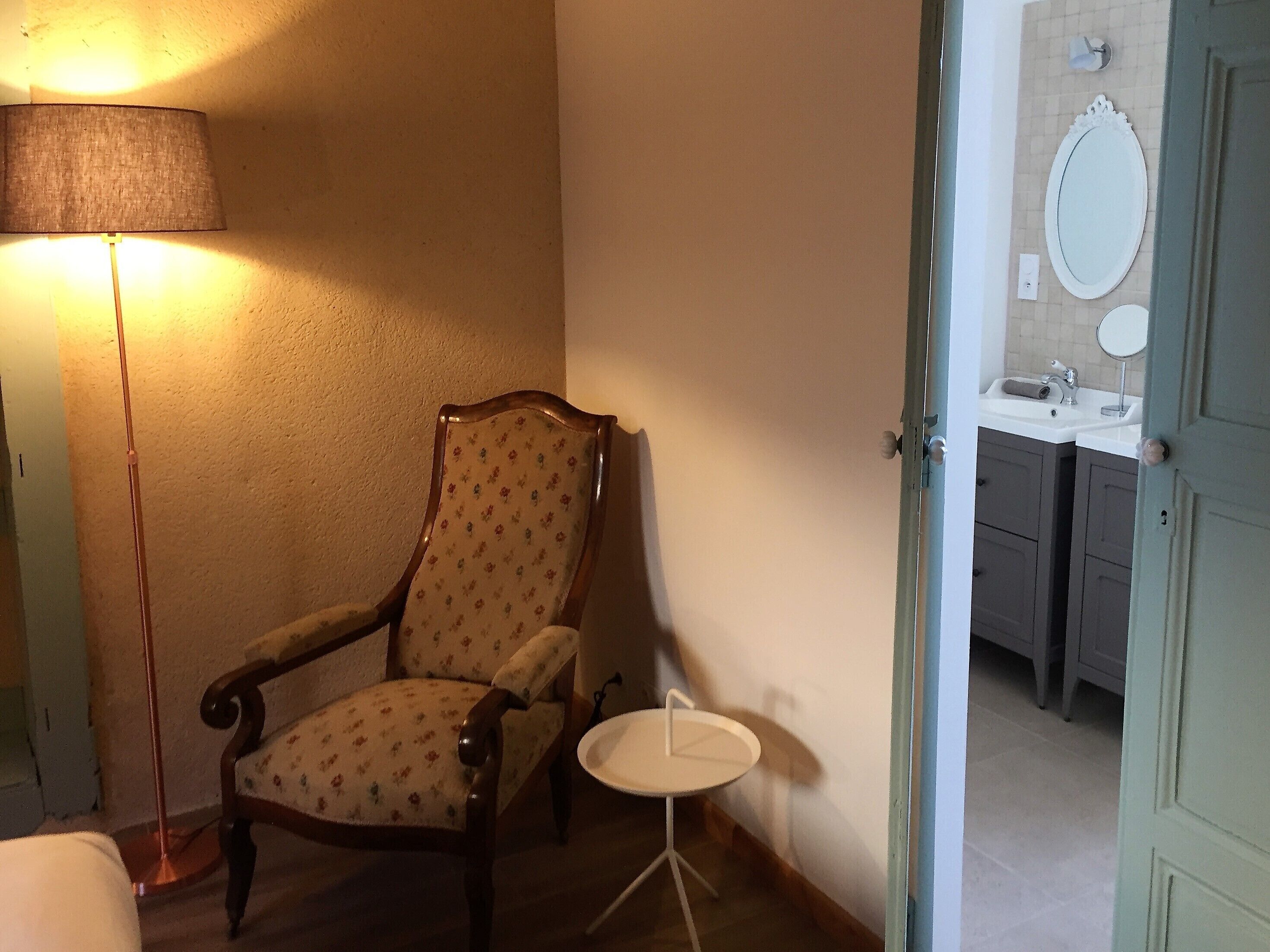 A room with a wooden chair, a small round table, a lamp, and a bathroom visible through an open door.