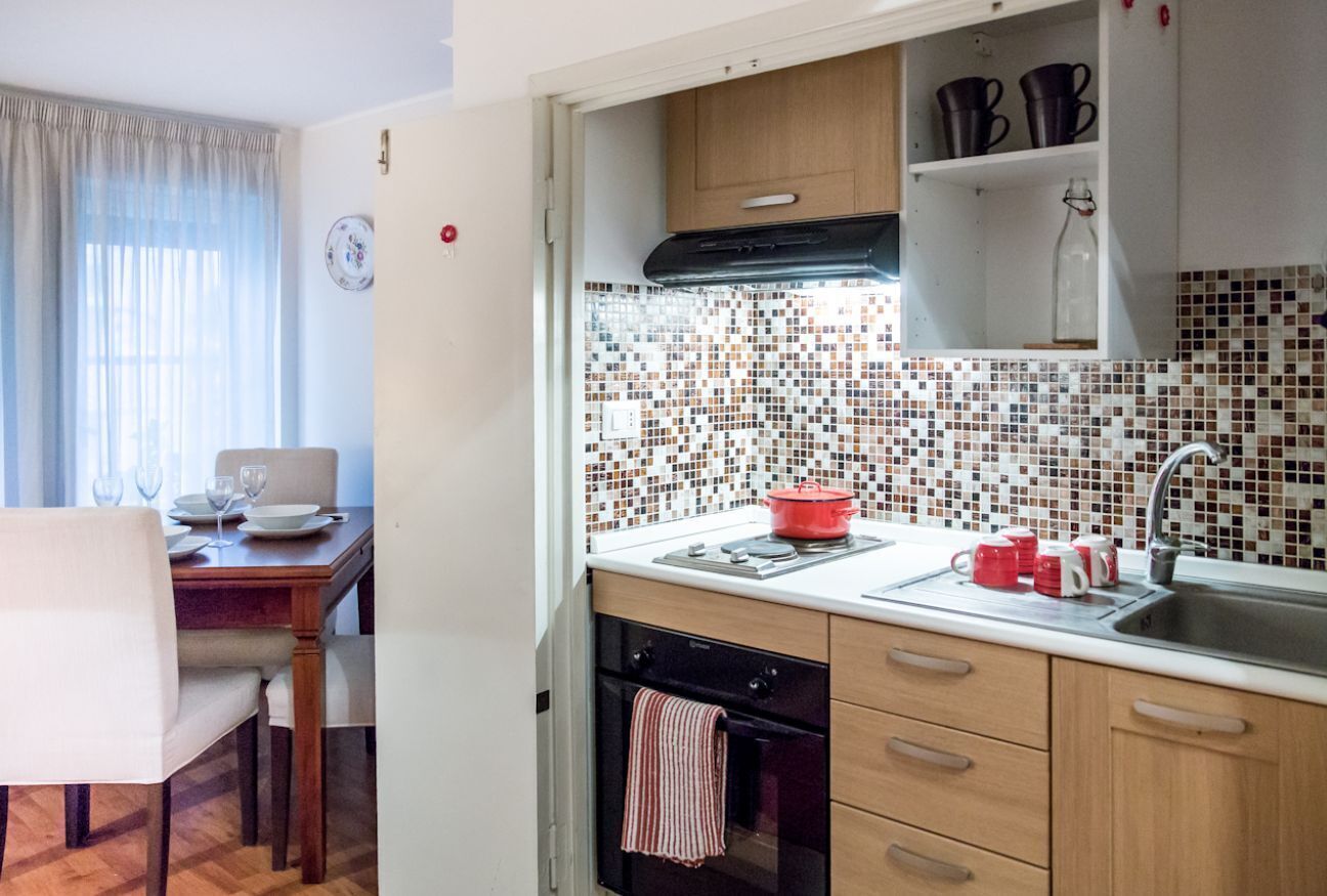 Una cucina compatta con rivestimento in piastrelle, mobili in legno, forno integrato e zona pranzo con tavolo e sedie.