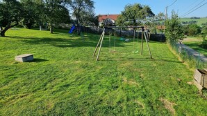 Un patio cubierto de césped con un columpio, un pequeño parque infantil y una casa de techo rojo al fondo.