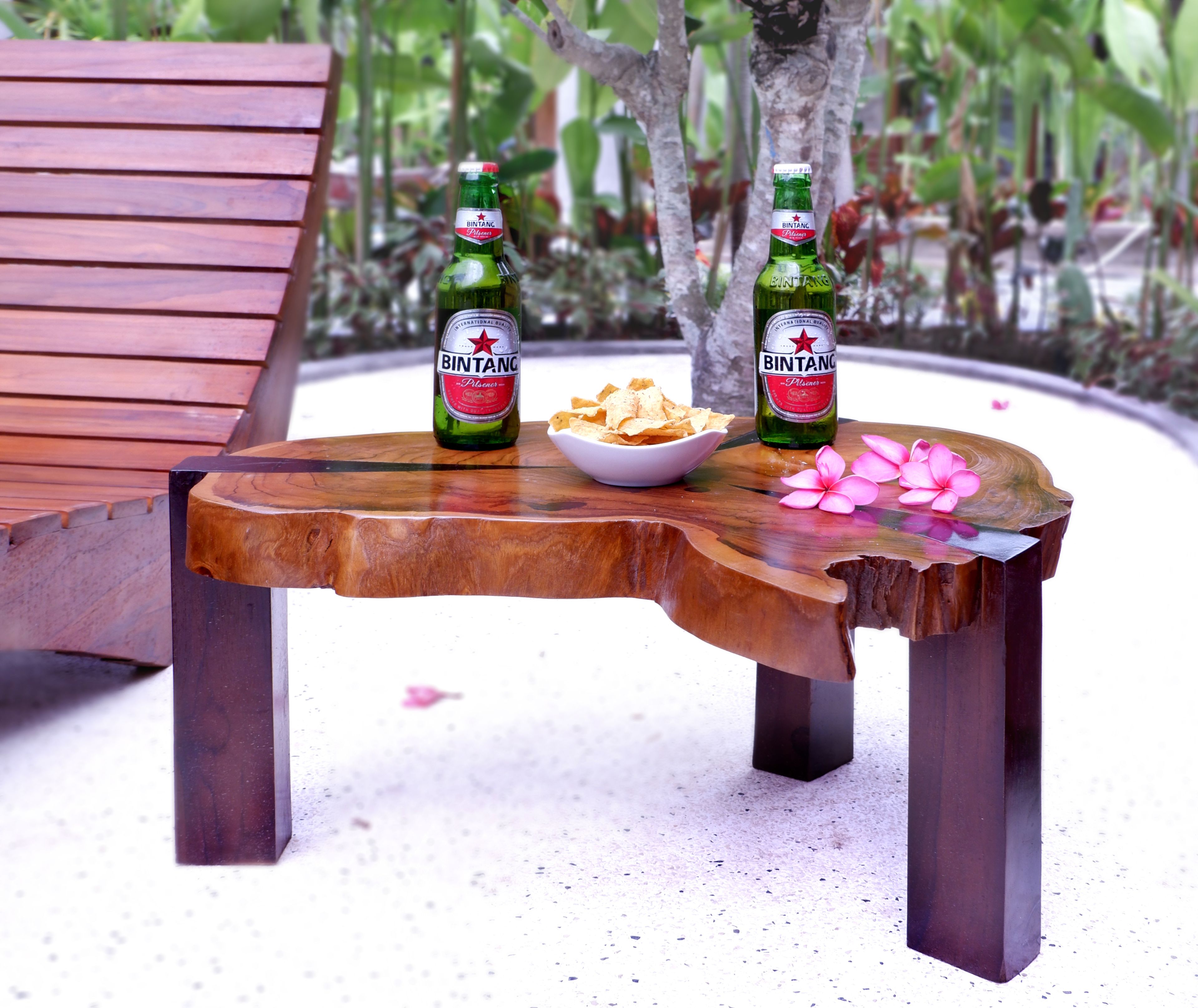 A wooden table with two bottles of Bintang beer, a bowl of chips, and pink flowers.