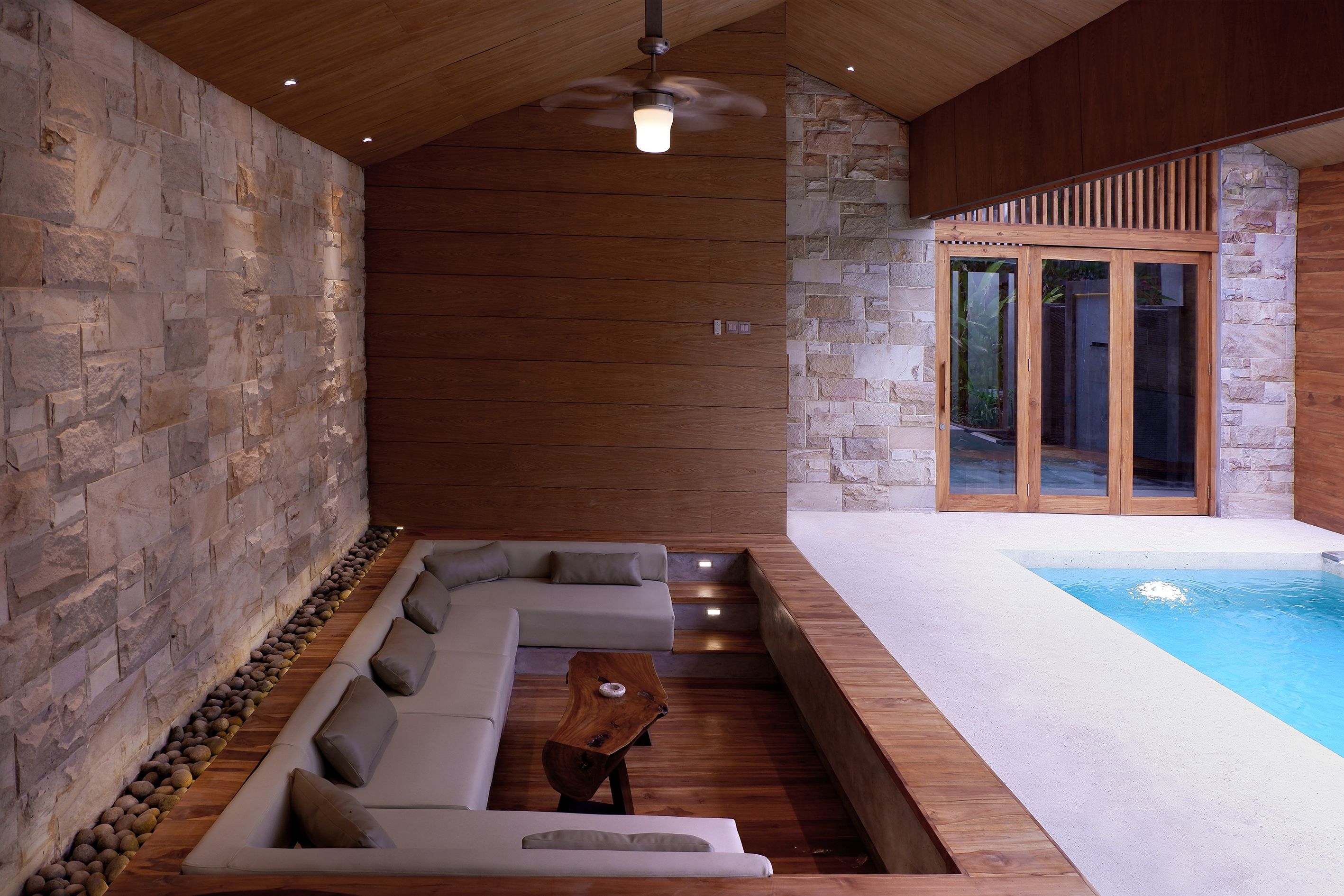 A modern indoor pool area with a stone wall, wooden ceiling, and a built-in seating area with cushions.