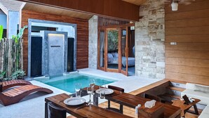 A modern outdoor pool area with a wooden deck, a dining table set for two, and a stone wall.