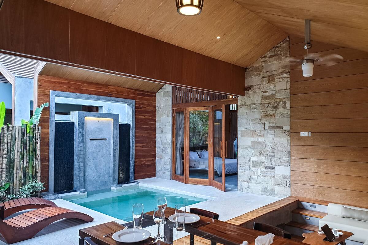 A modern outdoor pool area with a wooden deck, a dining table set for two, and a stone wall.