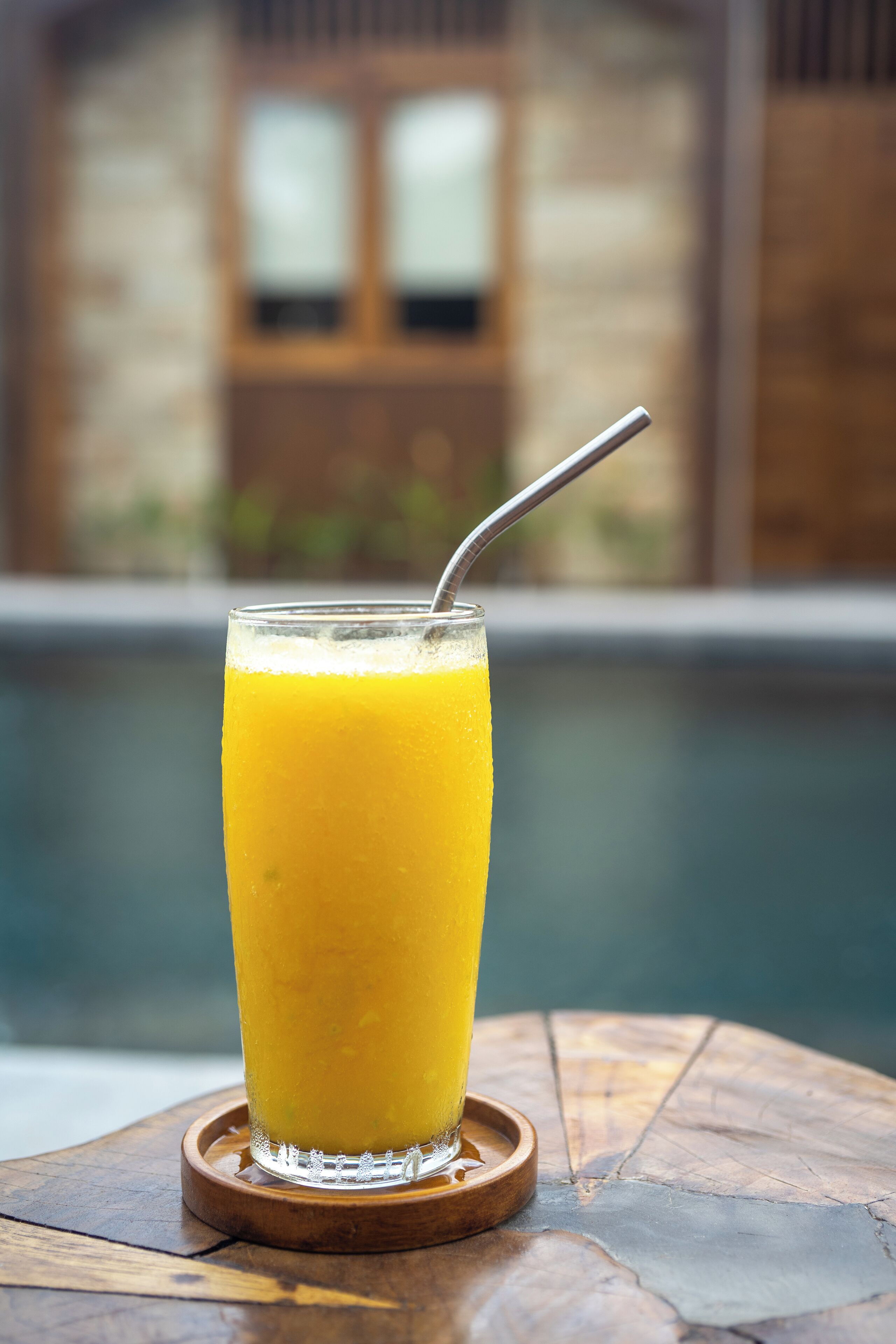 A tall glass of orange juice with a straw on a wooden coaster by a pool.
