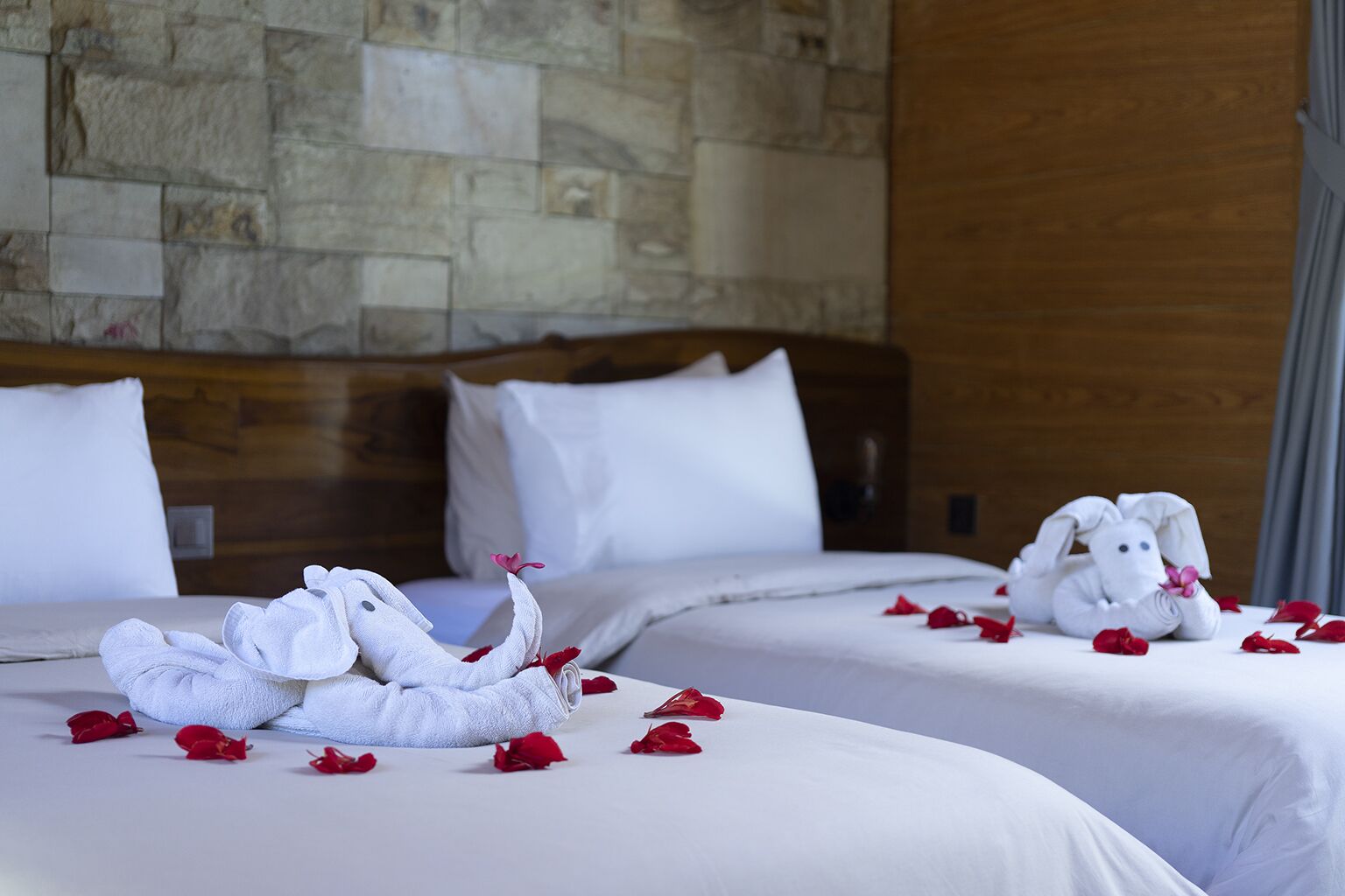 A hotel room with two beds, stone wall, wooden headboard, white bedding, and towels shaped like animals.