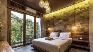 A modern bedroom with a large bed, wooden headboard, stone wall, and a view of greenery through large windows.