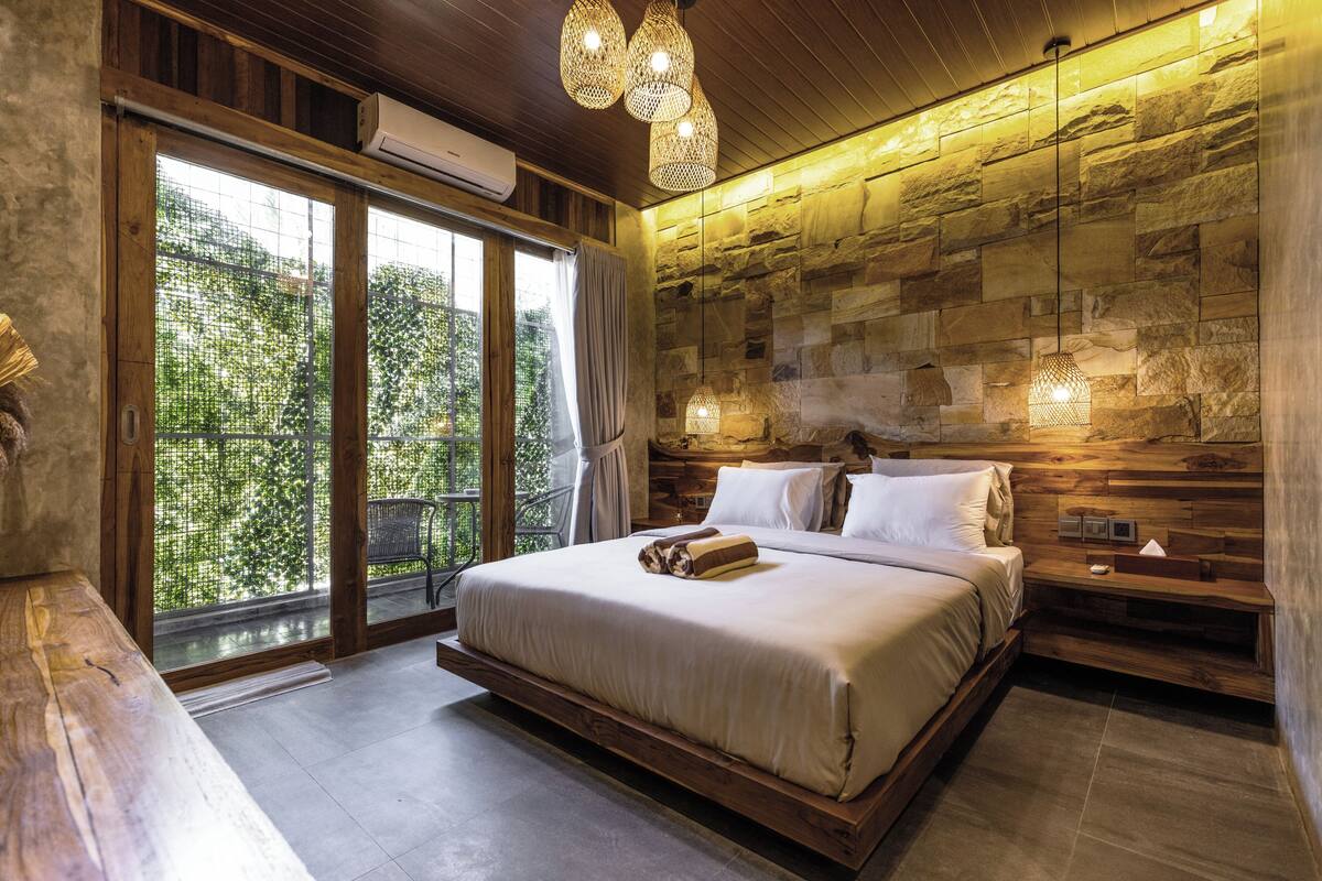 A modern bedroom with a large bed, wooden headboard, stone wall, and a view of greenery through large windows.