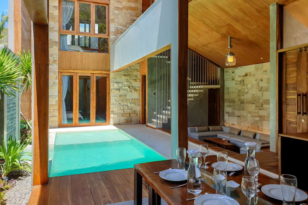 A modern outdoor dining area with a pool, wooden furniture, and stone walls.