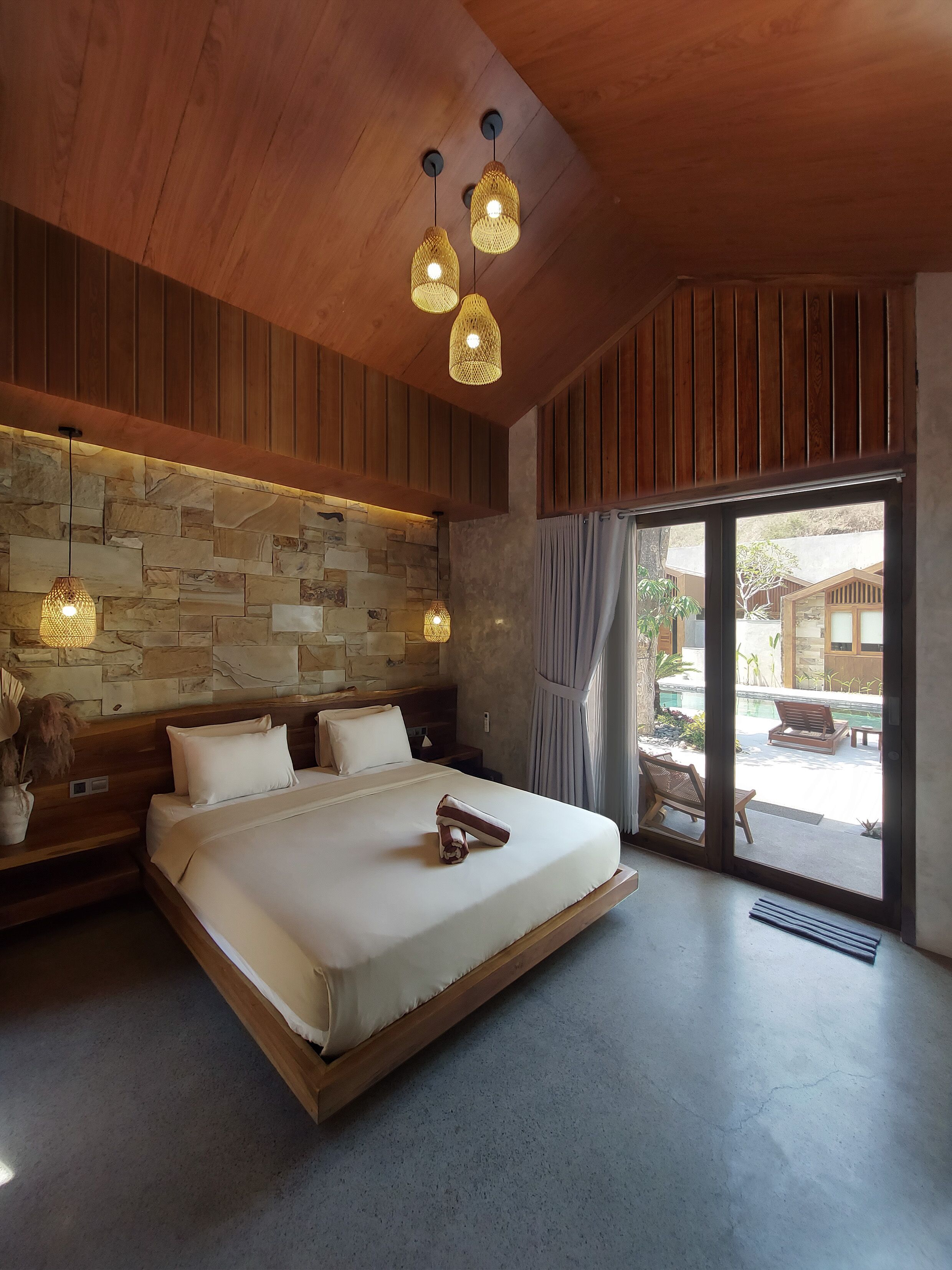 A modern bedroom with a stone wall, wooden ceiling, and a large bed with a wooden headboard. There are two pendant lights and a view of the outside through a glass door.