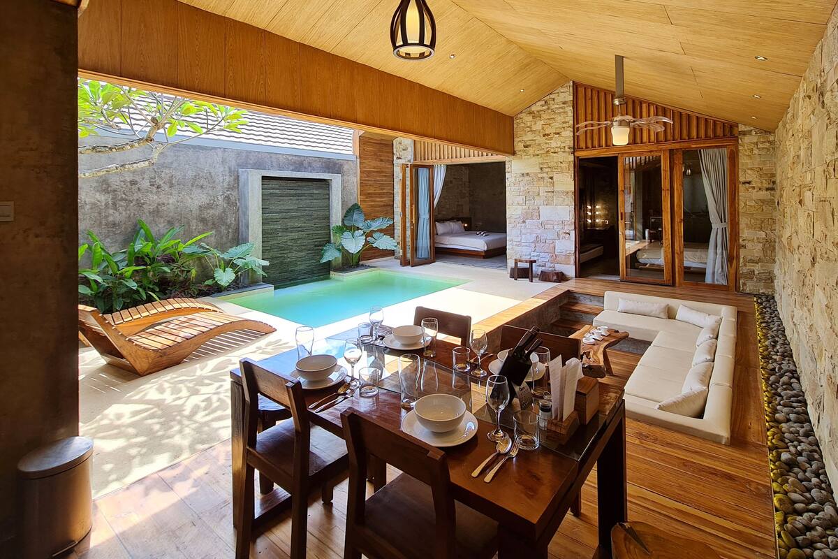 A modern outdoor dining area with a pool, wooden furniture, and a stone wall.