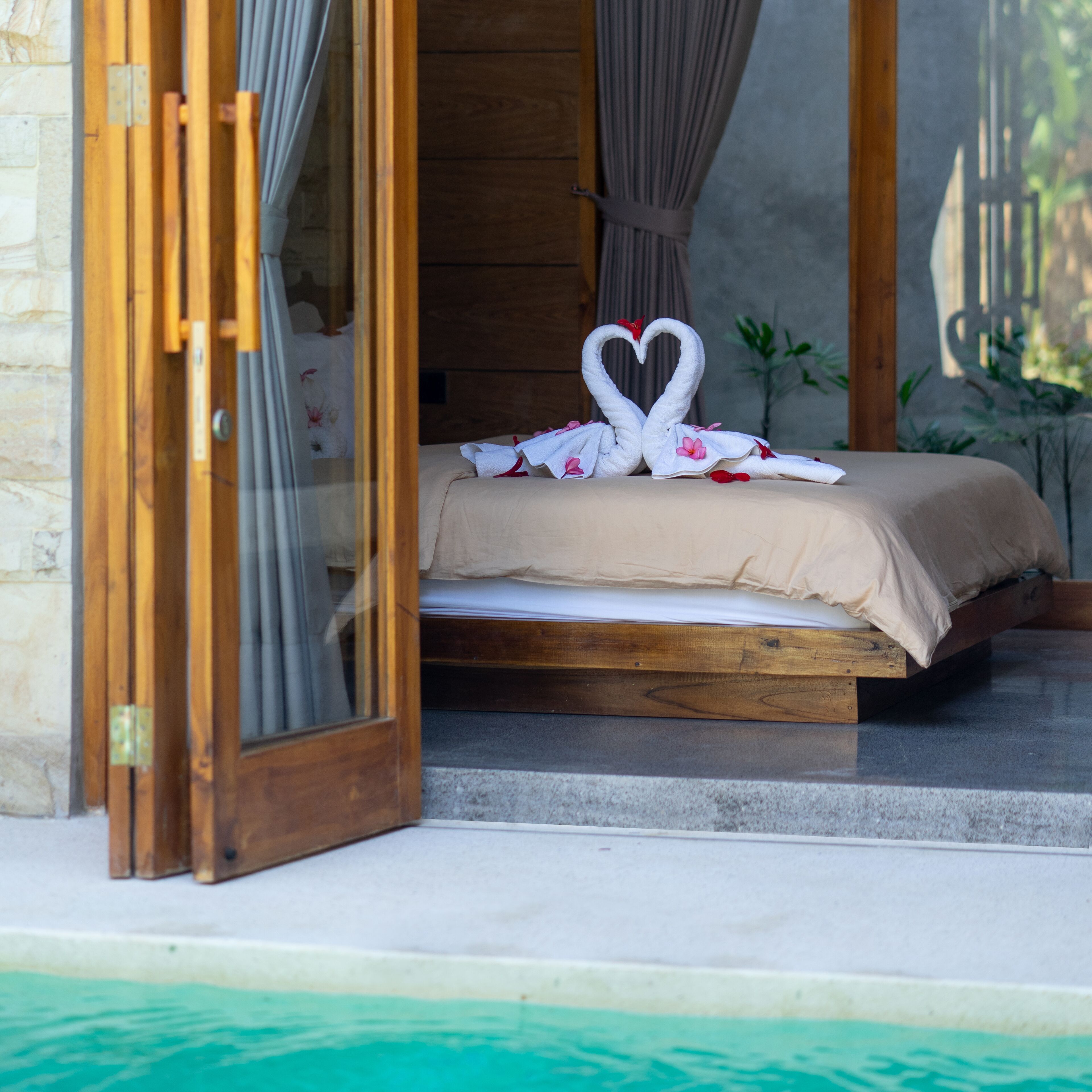 A bed with a heart-shaped towel decoration, a wooden headboard, and a view of a swimming pool.