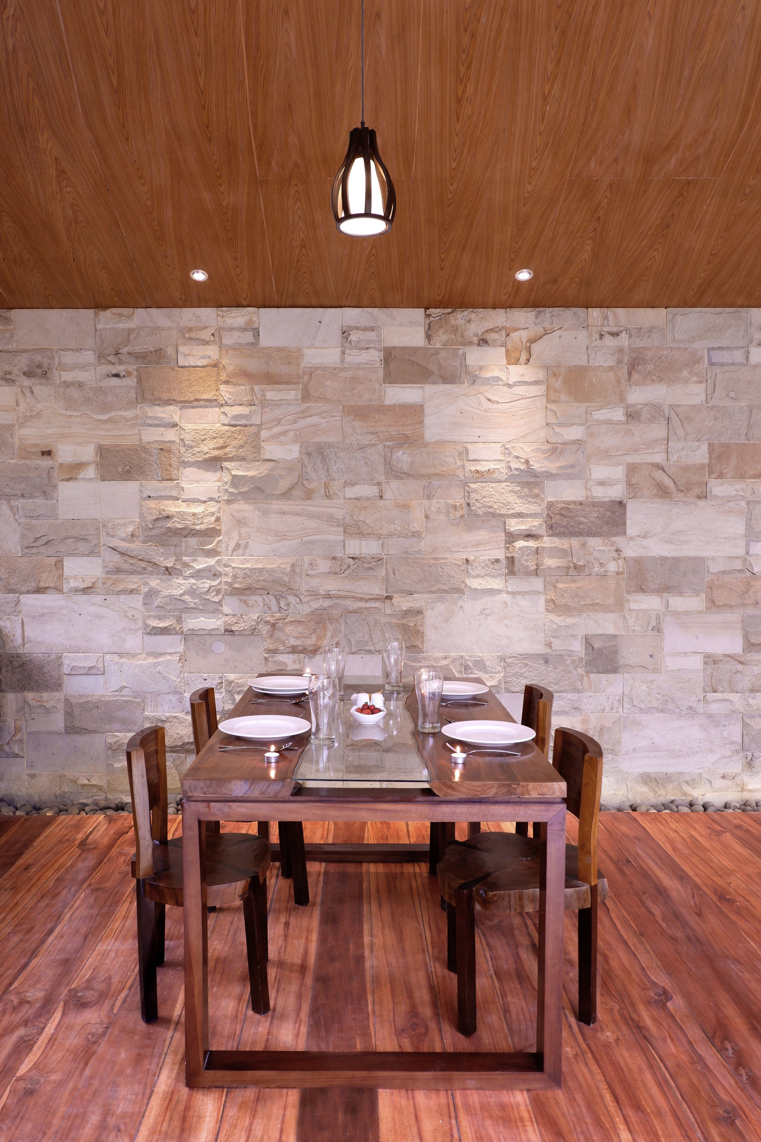 A dining area with a wooden table set for four, surrounded by chairs, against a stone wall backdrop.