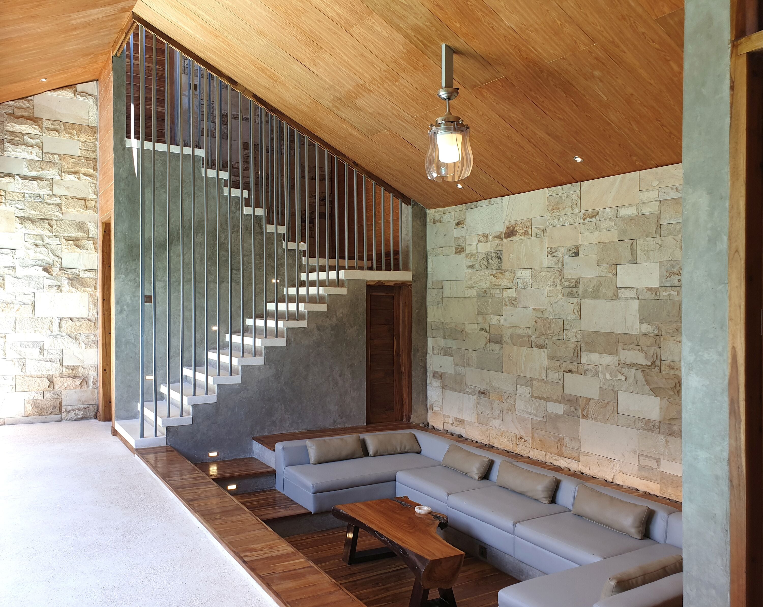 A modern interior with a wooden ceiling, a stone wall, a staircase with metal railings, and a light-colored sectional sofa.