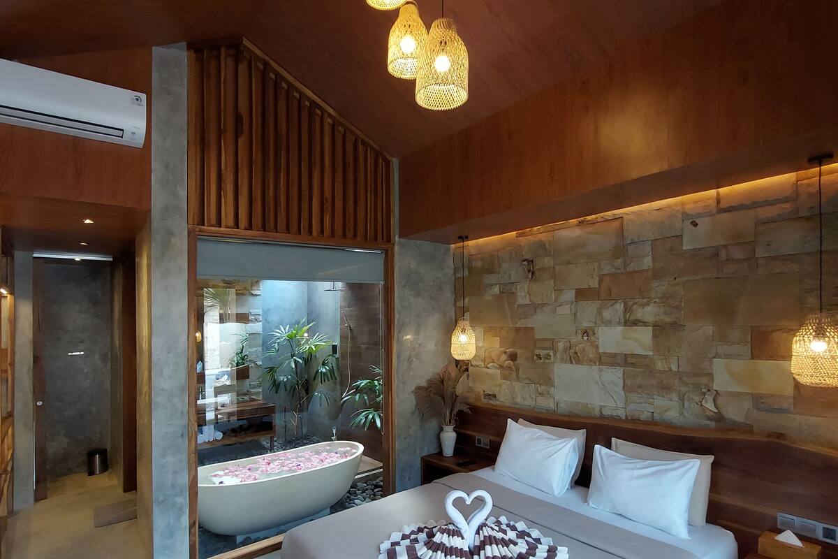 A modern bedroom with a large bed, wooden headboard, stone wall, and a view of a bathroom with a bathtub and plants.