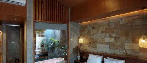 Ein modernes Schlafzimmer mit einem großen Bett, einem Holzkopfende, einer Steinwand und Blick auf ein Badezimmer mit Badewanne und Pflanzen.