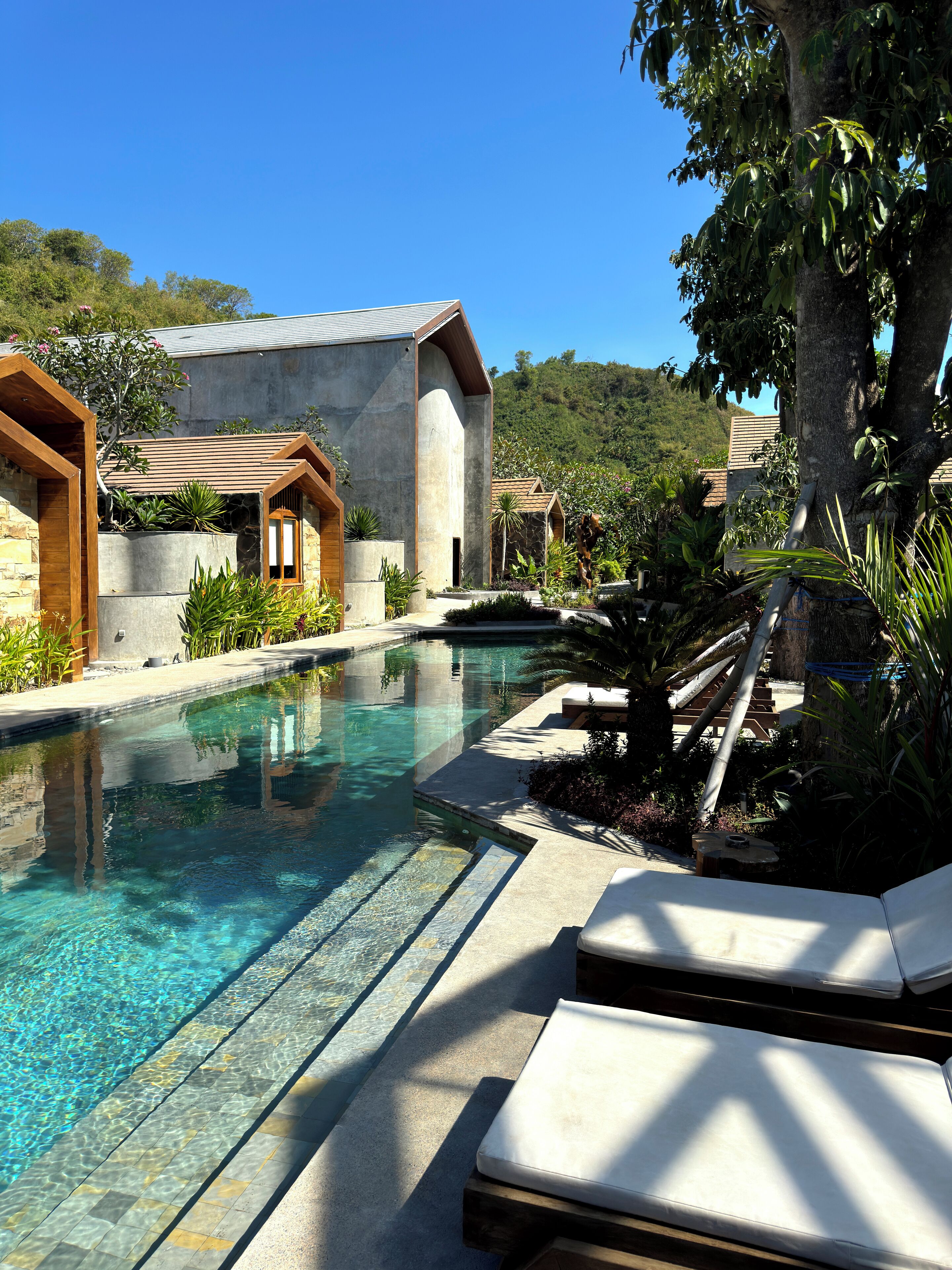 A pool area with clear water, lounge chairs, and a stone building with wooden accents.