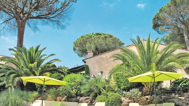A pool area with lounge chairs and umbrellas, surrounded by greenery and a building with a stone facade.