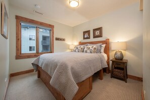 A bedroom with a large bed, a window with a view of a building, a bedside table with a lamp, and a small wooden nightstand.