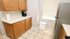 A compact kitchen with wooden cabinets, a countertop, a coffee maker, a trash bin, a window with blinds, and a refrigerator.