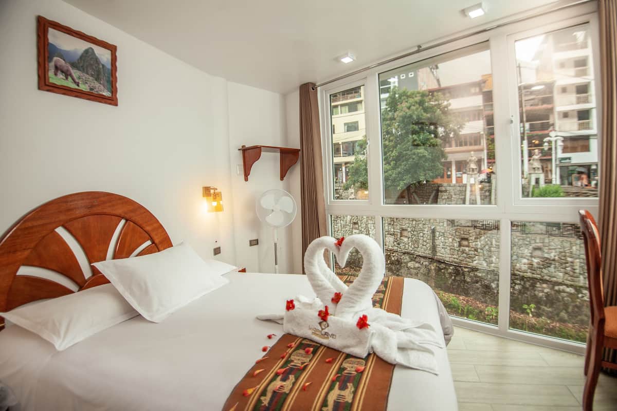 Habitación de hotel con un ventanal grande, cama con ropa blanca, cabecera de madera y una decoración de toallas.