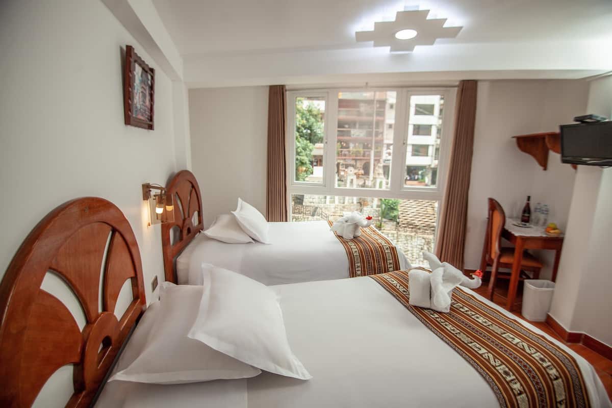 Habitación de hotel con dos camas, un escritorio, una silla, un televisor y ventana con vista a edificios.