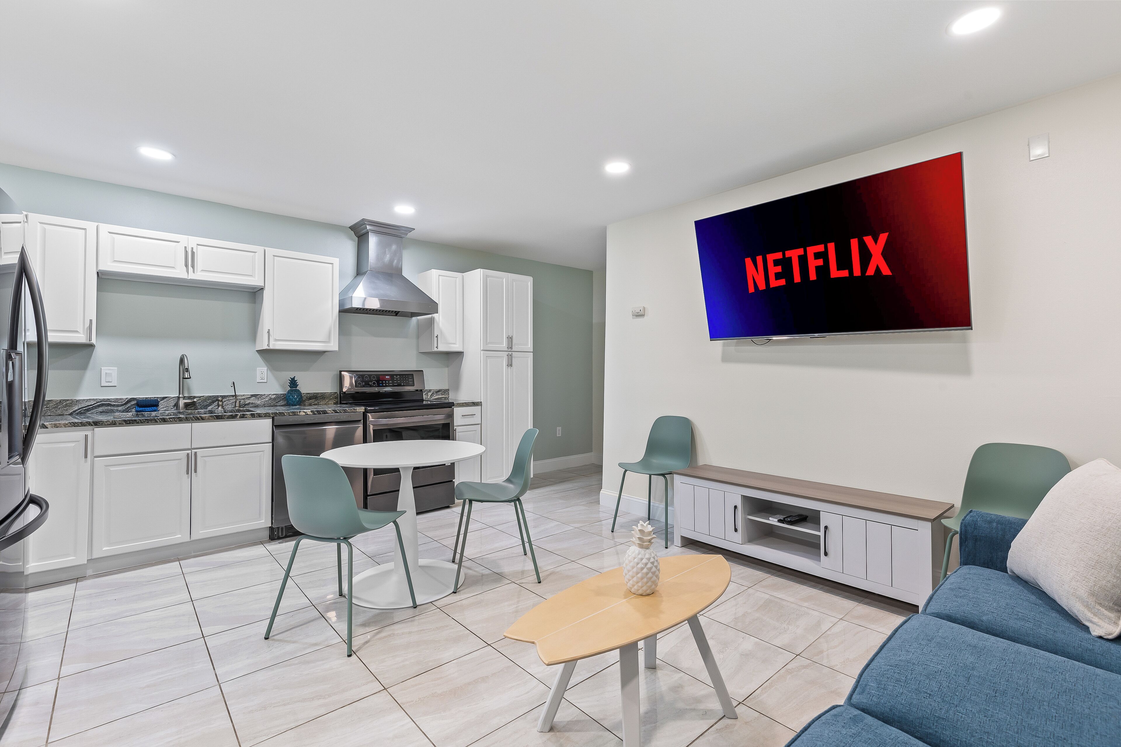 Een moderne keuken met witte kasten, een roestvrijstalen afzuigkap en een eethoek met een ronde tafel en stoelen. Een flatscreen televisie aan de muur toont het Netflix logo.