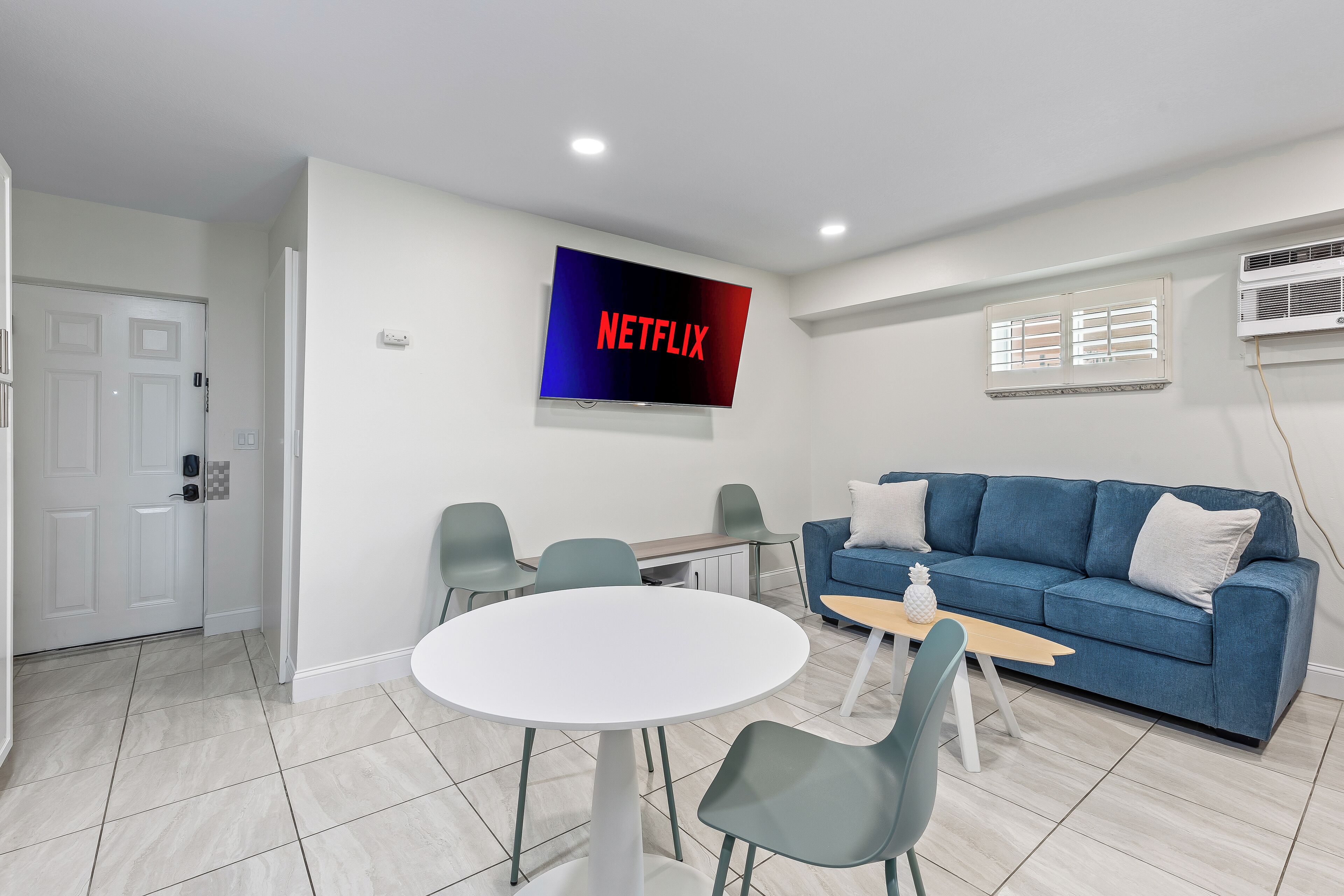 A modern living room with a flat-screen TV displaying "NETFLIX," a blue sofa, a round white table, and four chairs.