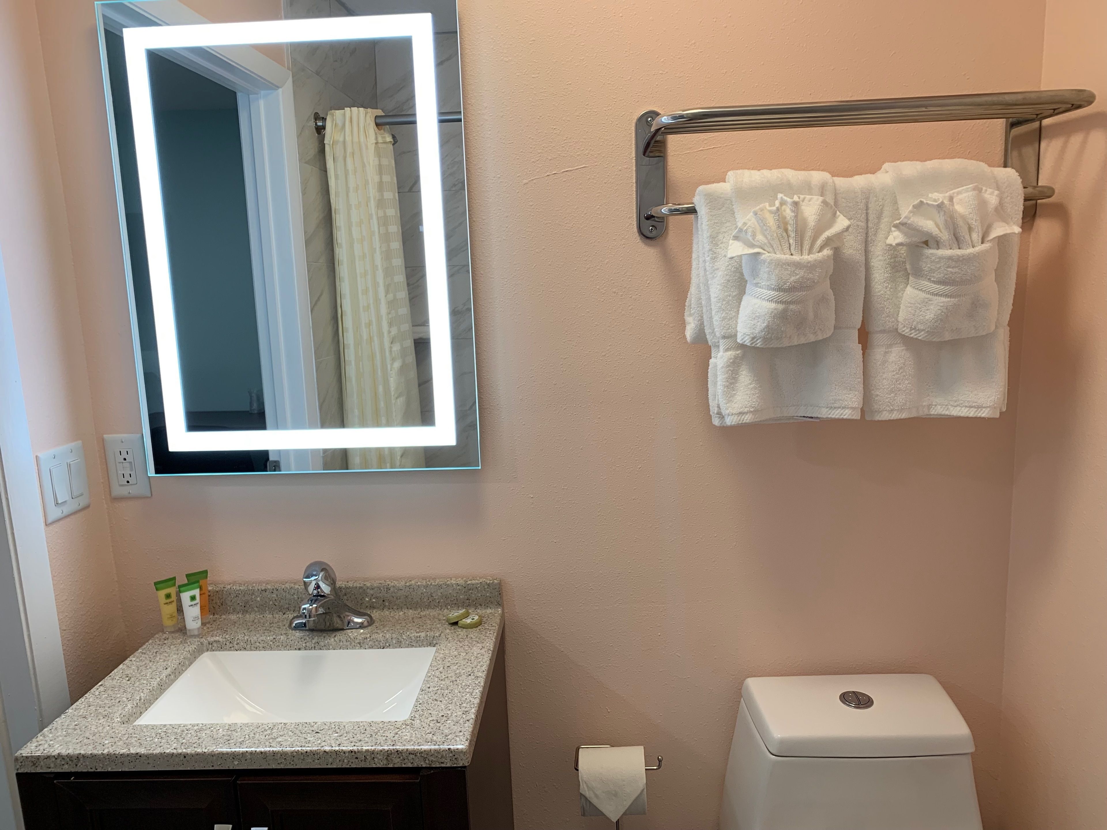 A bathroom with a granite sink, a mirror with a light, a towel rack with two towels, a toilet, and a shower curtain.