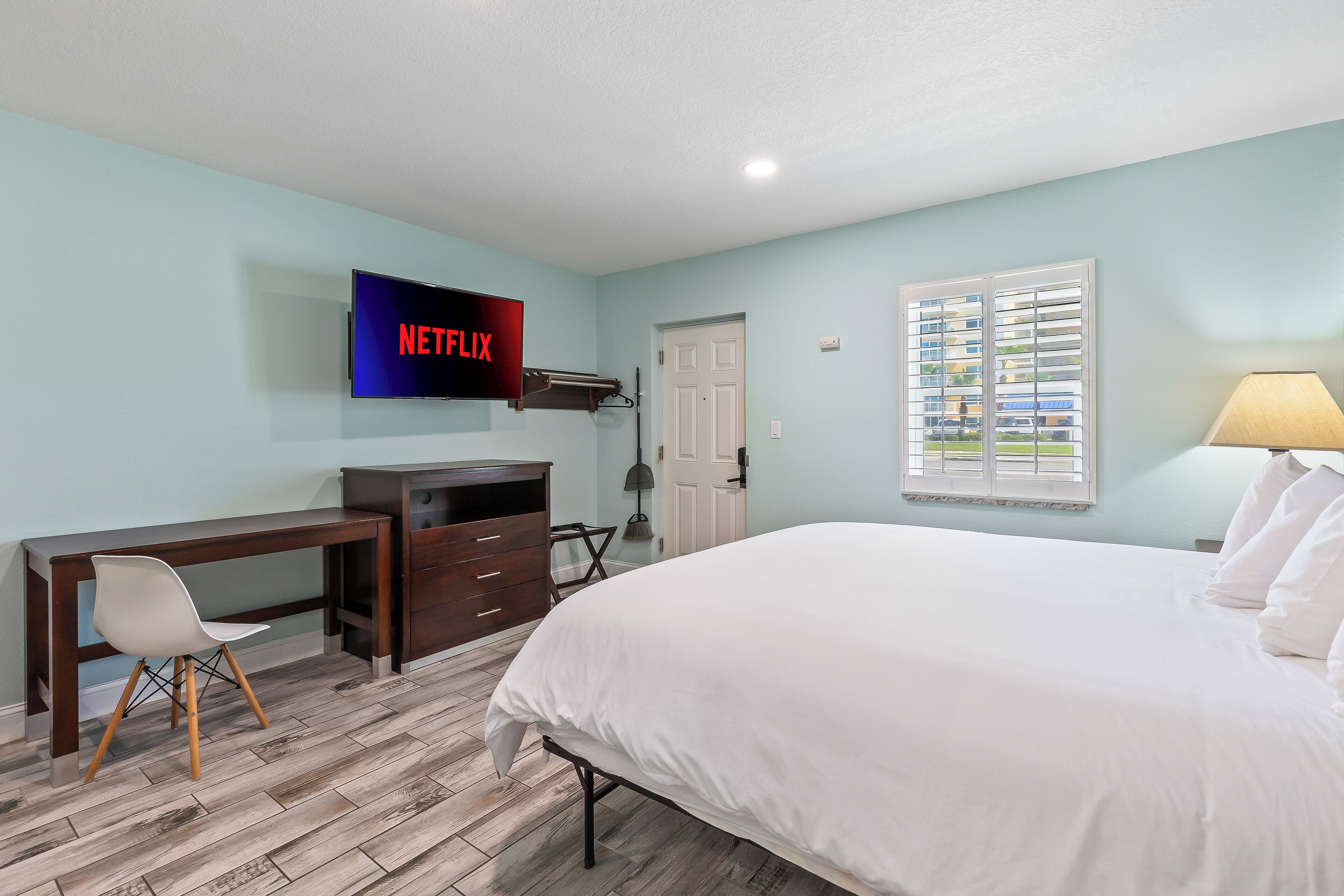 A bedroom with a bed, a desk, a chair, a TV, and a window with blinds.