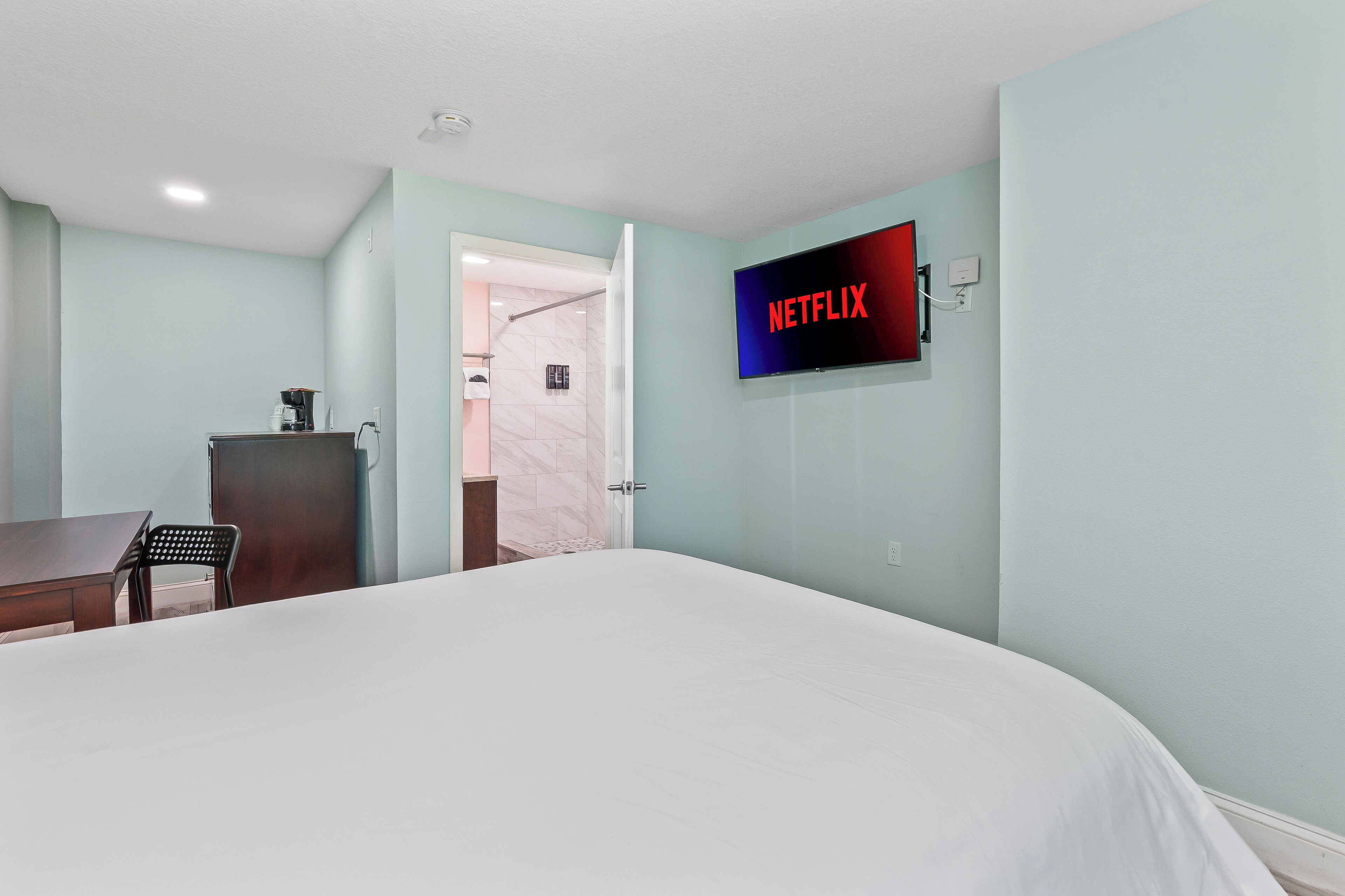 A hotel room with a bed, a desk, a chair, a TV mounted on the wall, and a door leading to a bathroom.